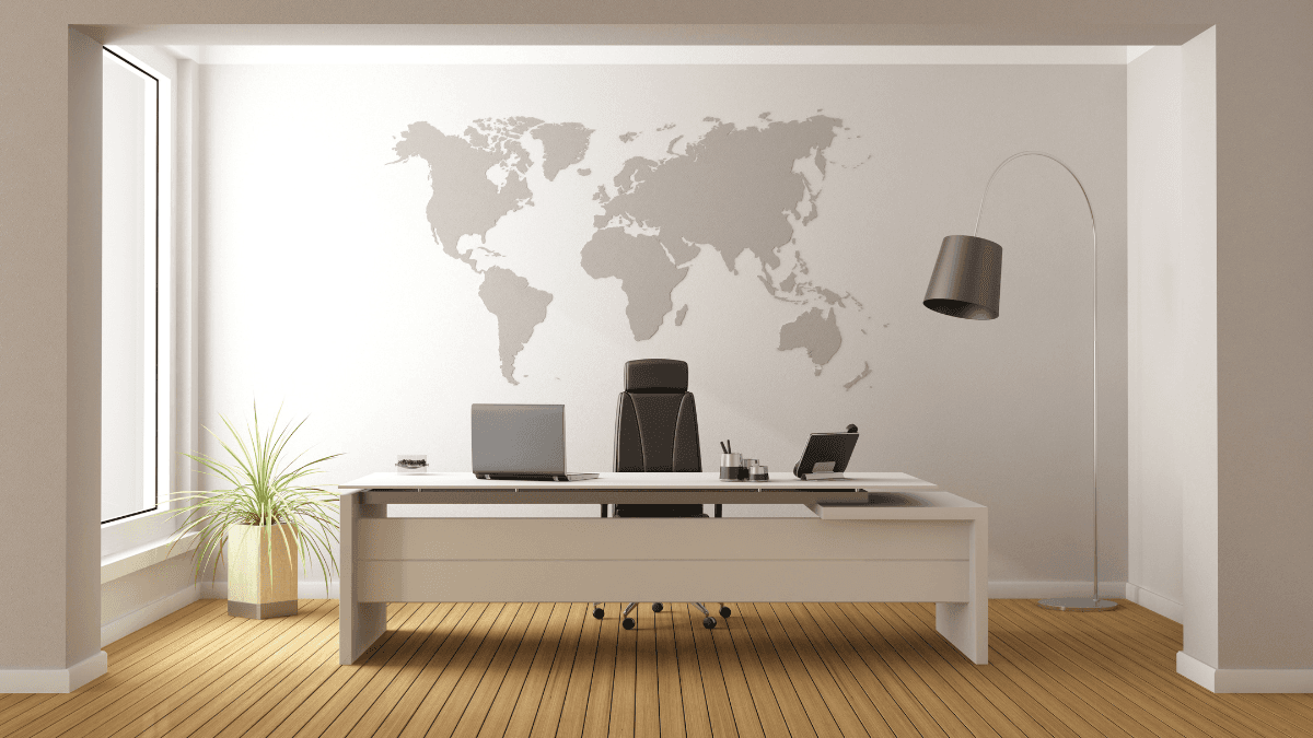 A modern office setting featuring a sleek white desk with a black chair, an open laptop, and minimalistic decor, all positioned against a wall adorned with a world map. The room has wooden flooring and a large window allowing natural light to enhance the space.