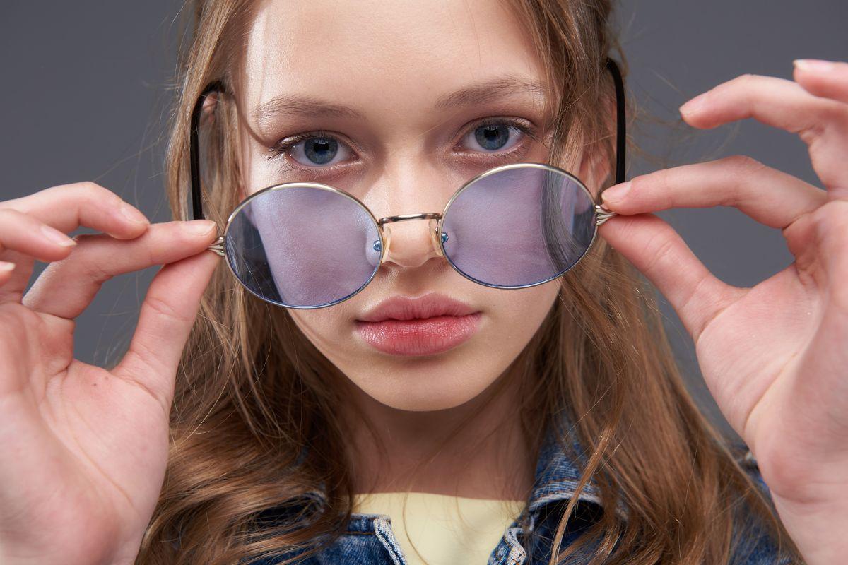 A close-up of a young woman adjusting her round sunglasses, gazing directly into the camera.
