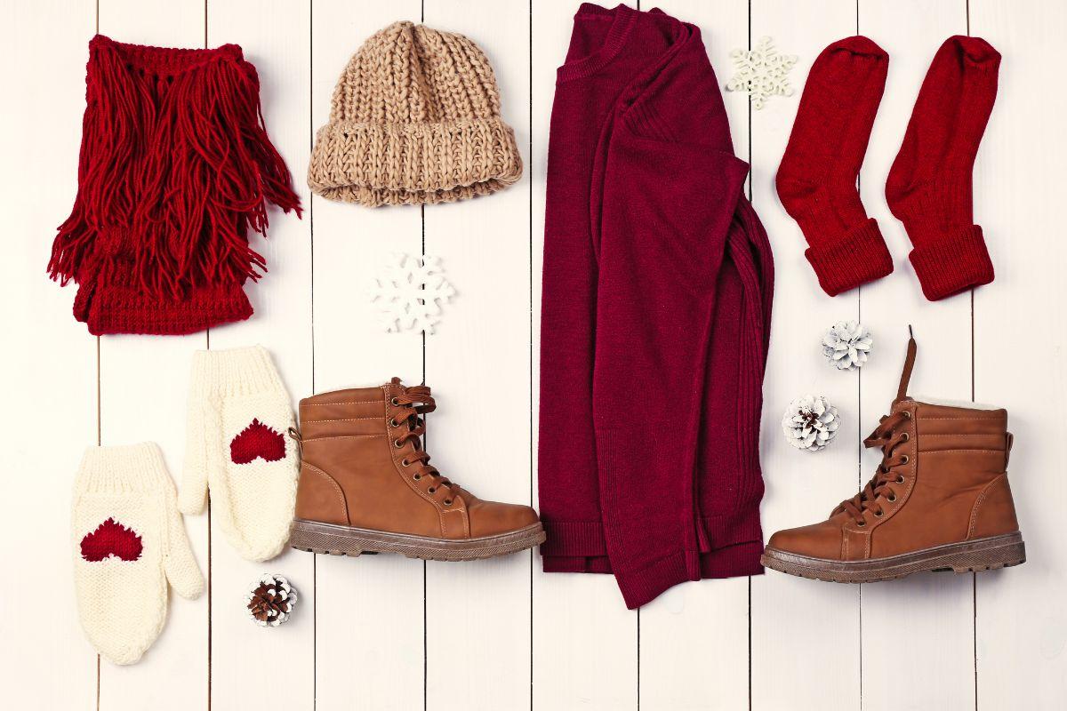 Flat lay of cozy winter clothing and accessories, including a red sweater, scarf, socks, beige hat, mittens with heart patterns, and brown boots, arranged on a white wooden background with snowflakes and pinecones.