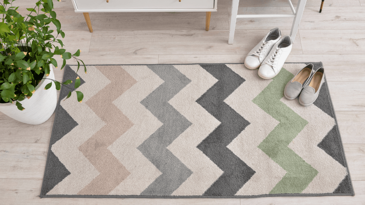 Chevron-patterned rug in earthy tones, placed on a light wood floor, accompanied by casual footwear and a potted plant, creating a welcoming entryway vibe.