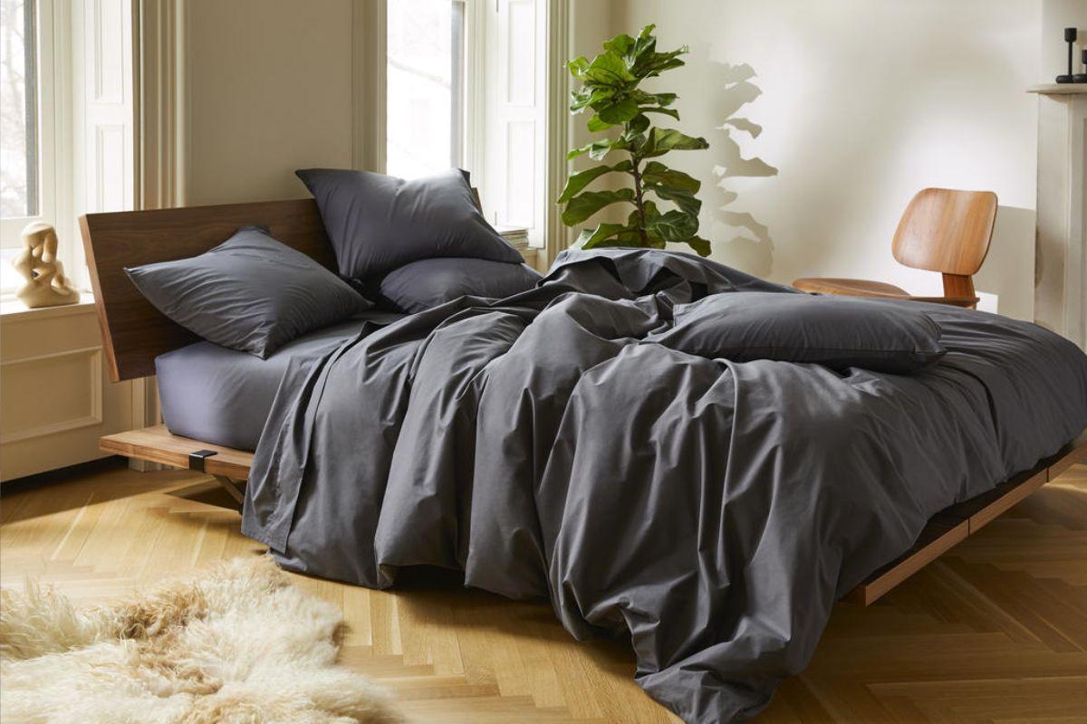 Modern bedroom with a neatly designed wooden bed frame, dressed in dark gray bedding, surrounded by natural light, a potted plant, and minimalist decor.