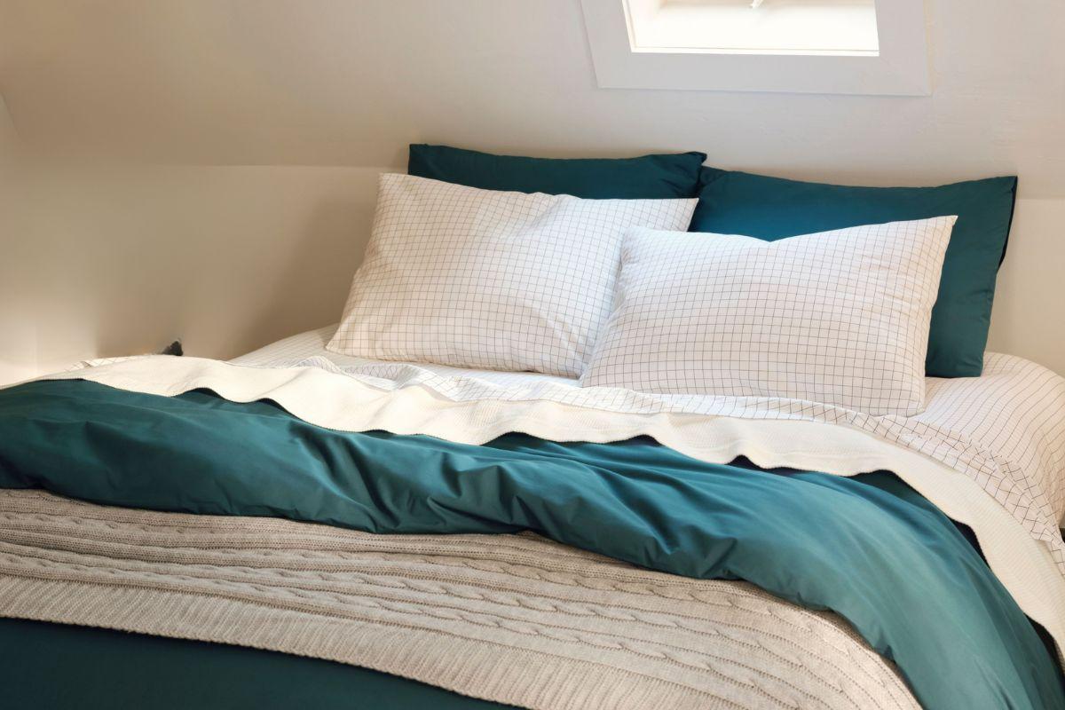 Inviting bed with teal bedding, white grid-patterned pillows, and a soft knitted throw under a small skylight.