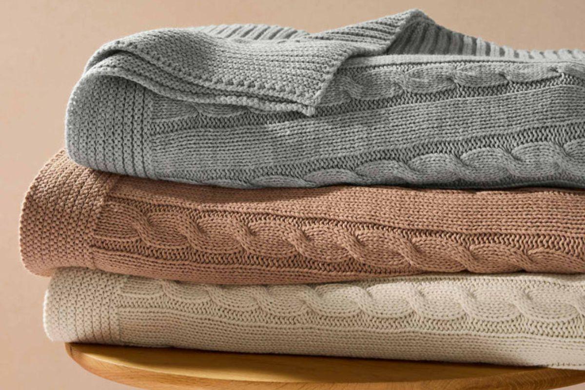 Stack of folded cable-knit blankets in gray, beige, and cream tones on a wooden surface.