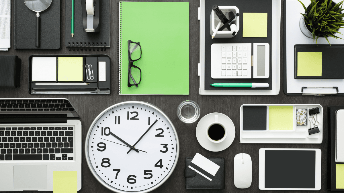Modern office desk setup featuring a large clock, laptop, tablet, notebooks, stationery, and a cup of coffee, accented with green sticky notes and office supplies for a clean and organized workspace.
