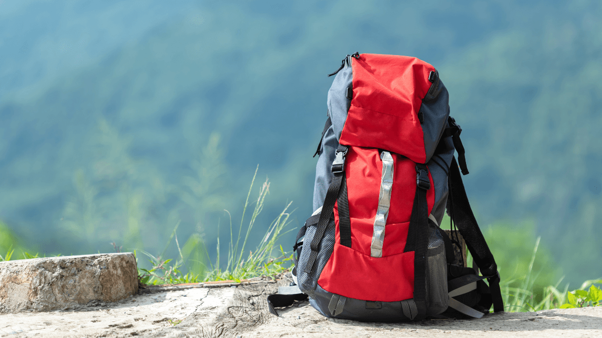 Red and black hiking backpack resting on a mountain trail with a scenic green landscape in the background.