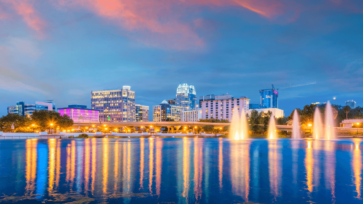 Orlando cityscape at twilight, with illuminated fountains reflecting vibrant hues on a serene lake.