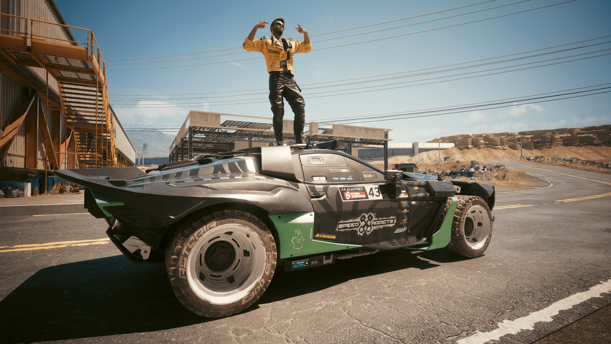 A man standing on a futuristic off-road car, posing with raised arms in an industrial desert setting under a clear blue sky.