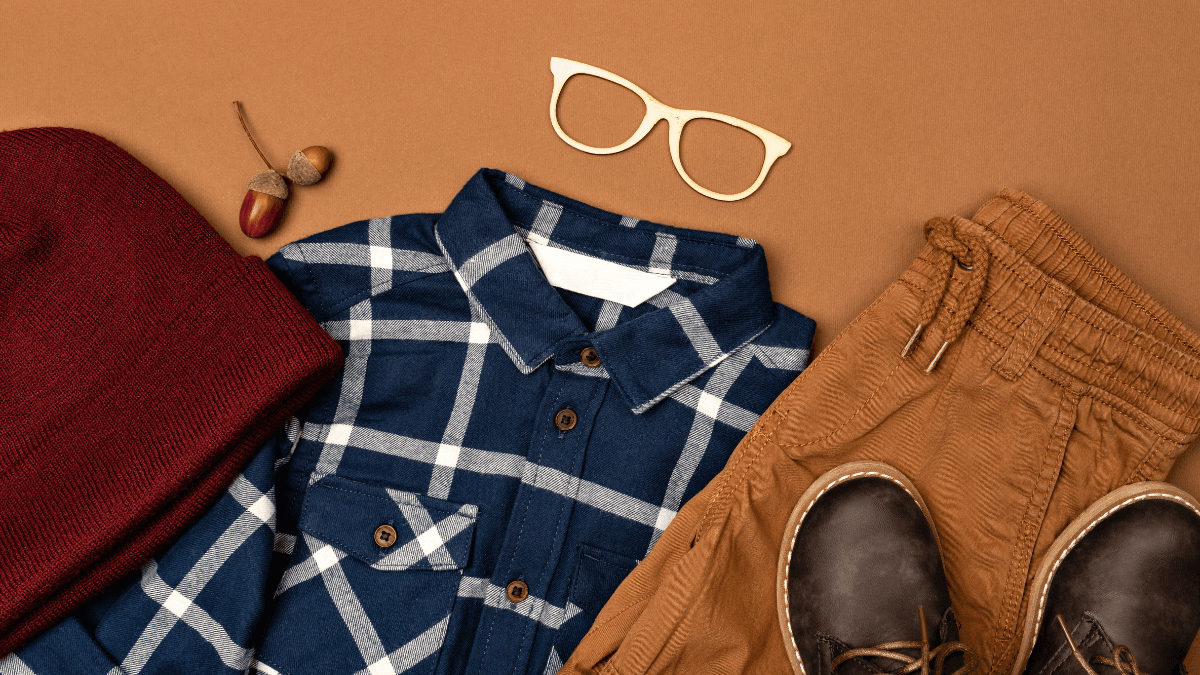 A flat lay of autumn-inspired clothing and accessories, featuring a blue plaid shirt, brown corduroy pants, leather boots, a maroon beanie, wooden glasses, and an acorn.