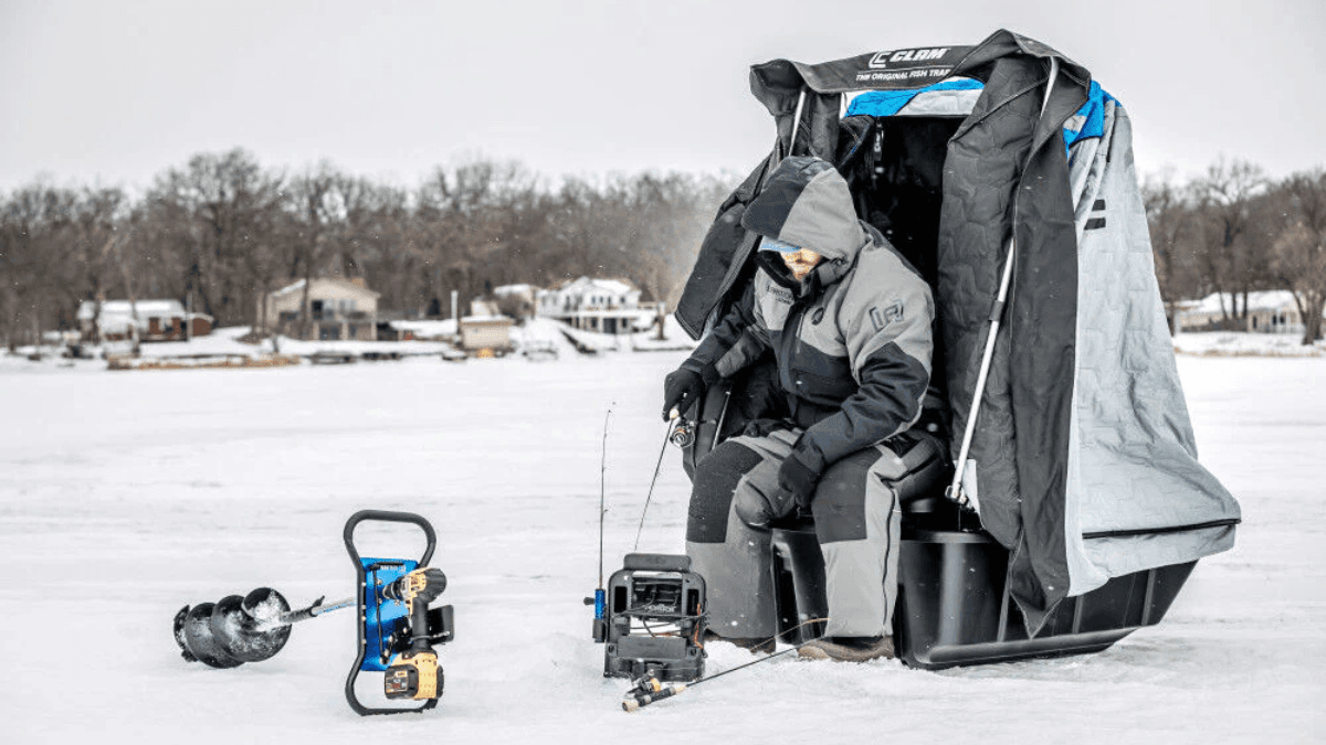 An ice fisherman sits inside a portable ice fishing shelter, dressed in heavy winter gear.