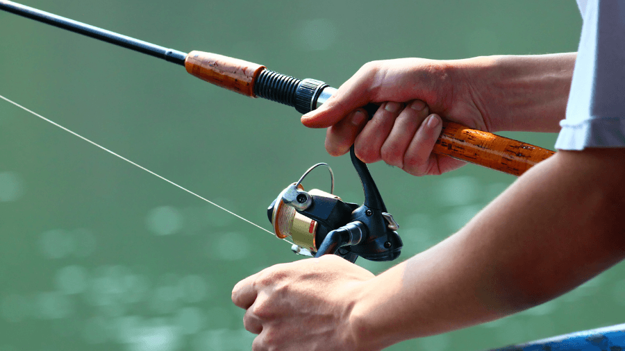 A close-up of a fisherman’s hands gripping a spinning reel fishing rod over a calm body of water.