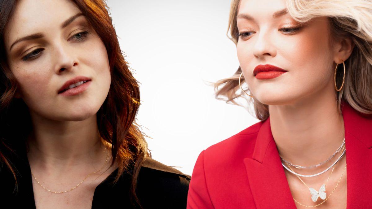 A close-up of two women wearing elegant gold jewelry, including delicate necklaces and hoop earrings, styled with black and red outfits.