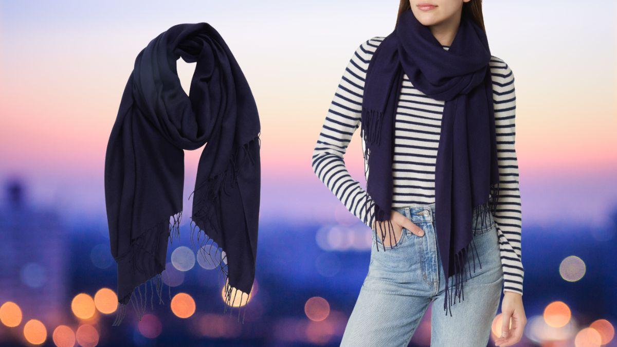 A navy blue cashmere and wool scarf is shown in two views—one neatly draped and the other worn casually over a striped top with jeans, against a blurred city skyline at sunset.