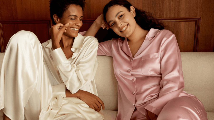 Two women lounging on a couch, wearing silky satin pajamas—one in cream and the other in blush pink—smiling and enjoying a relaxed moment together.