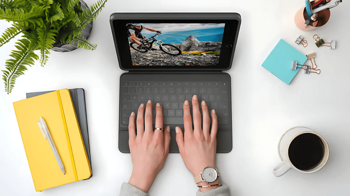 Top view of a person typing on a tablet with a detachable keyboard, surrounded by coffee, notebooks, and office supplies