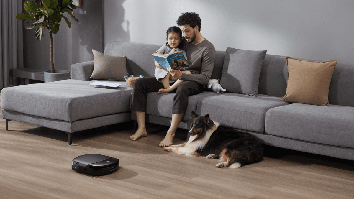Father and daughter reading on a gray sofa while a smart robot vacuum cleans the floor near their dog.
