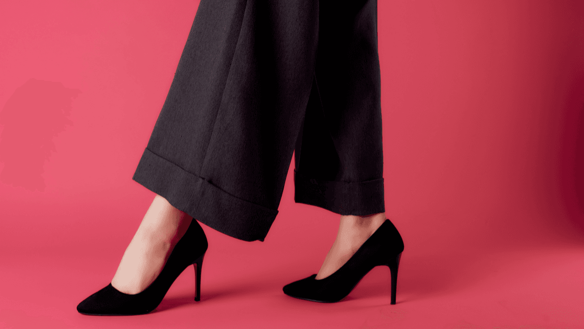 Person wearing black high heels and dark wide-leg trousers, walking against a pink background.