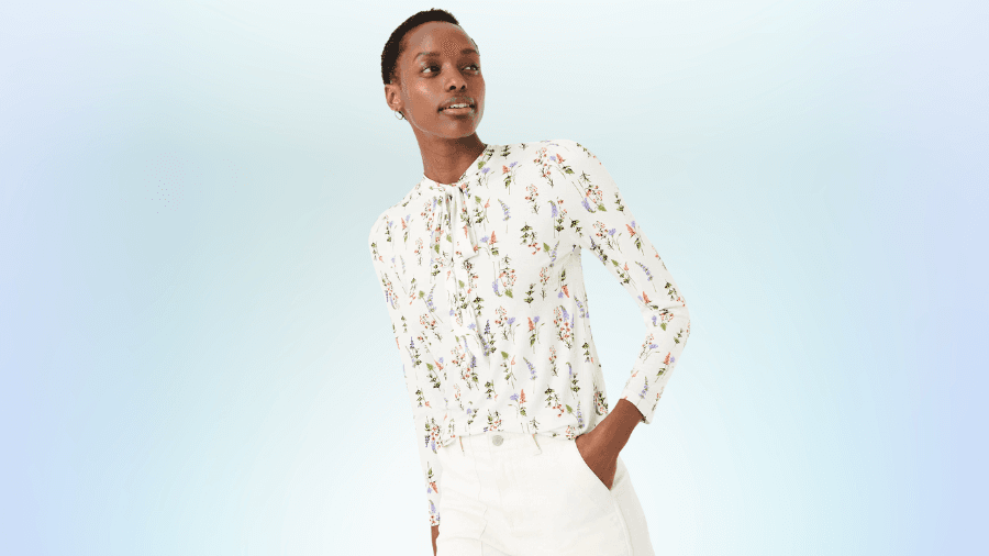 Woman wearing a white long-sleeve floral blouse tucked into white pants, posing confidently against a light gradient background.