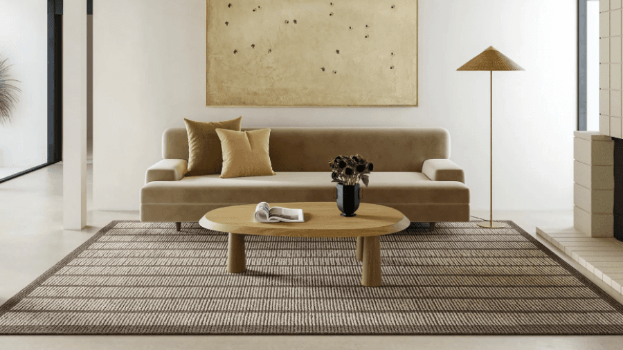 Modern living room with a beige sofa, green and mustard accent pillows, a wooden oval coffee table, and a striped area rug, complemented by a floor lamp with a woven shade.