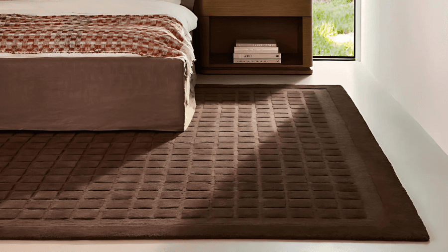 Modern bedroom featuring a brown bed with a textured red and white blanket, a matching brown nightstand with stacked books, and a dark brown rug with a square grid pattern on a light floor.