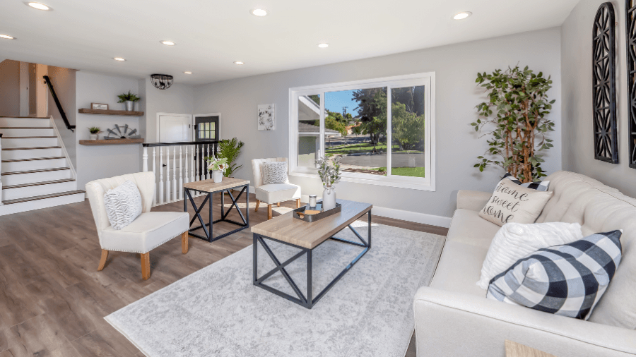 Bright, modern living room with light gray walls, large window, beige sofa with patterned pillows, two white armchairs, a light gray area rug, and wooden coffee tables with black metal frames.
