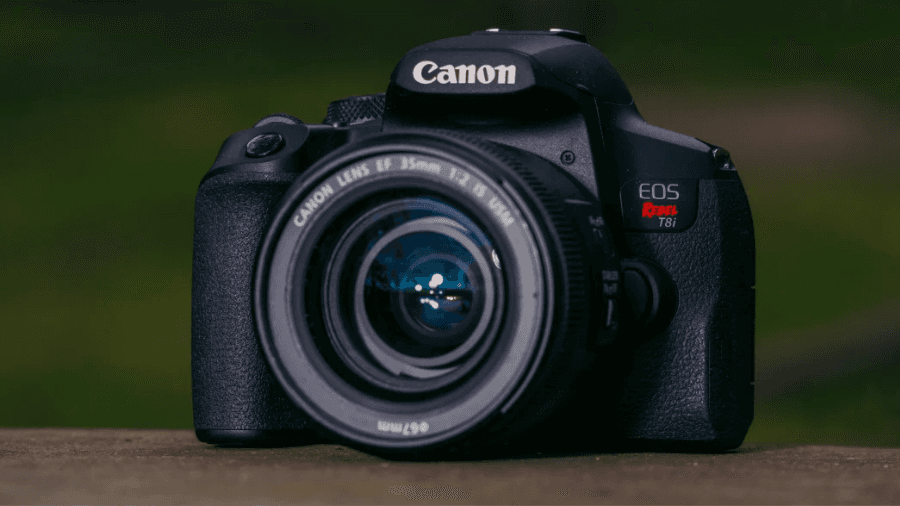 A Canon EOS Rebel T8i DSLR camera with an EF 35mm f/2 IS USM lens, placed on a wooden surface with a blurred green background.