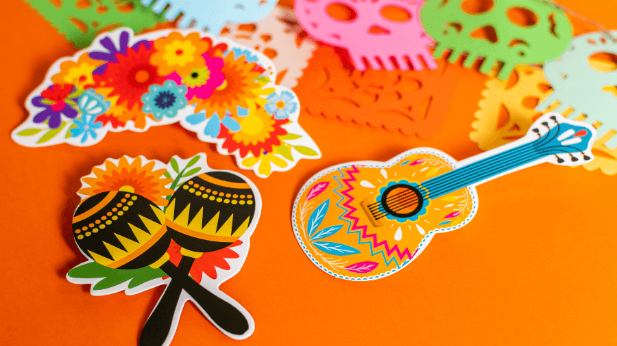 Colorful Día de los Muertos decorations featuring vibrant maracas, a decorated guitar, flowers, and papel picado skull designs on an orange background.