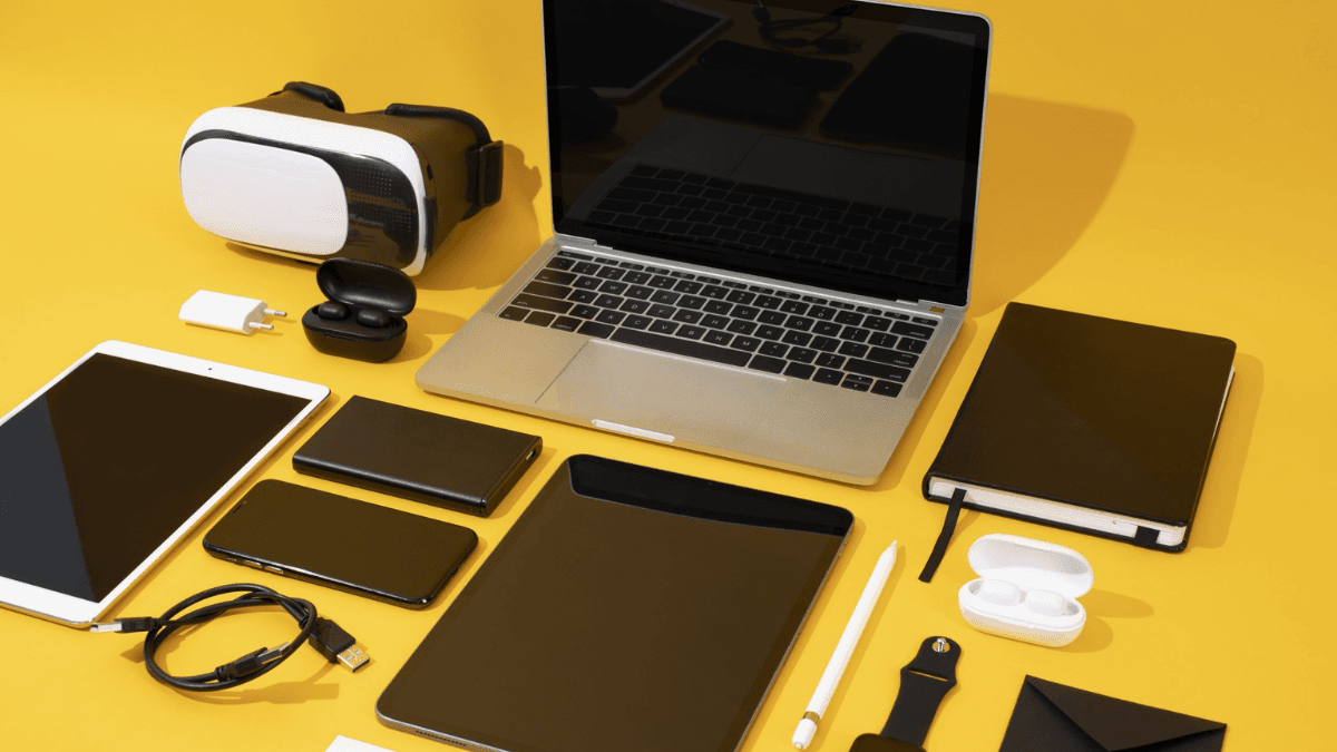 Collection of modern tech gadgets including a laptop, tablet, smartphone, smartwatch, VR headset, earbuds, and accessories on a yellow background.