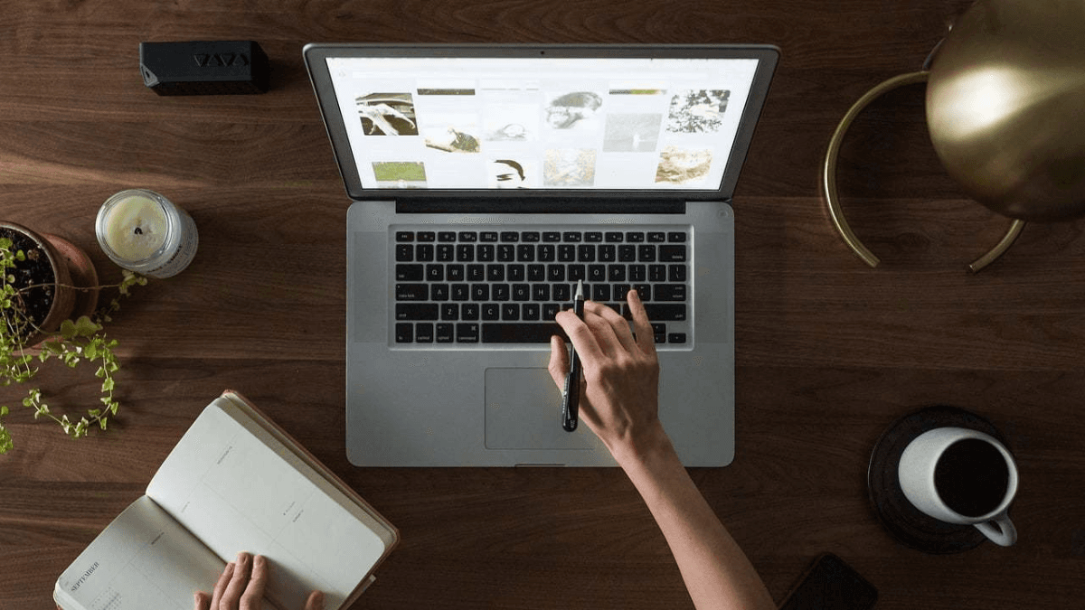 Person working on a laptop at a wooden desk with an open notebook, coffee cup, and plant.