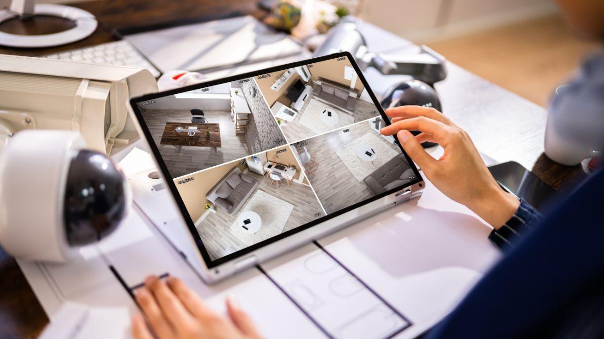 A person monitors a home security system on a tablet, showing four live camera feeds of different rooms.