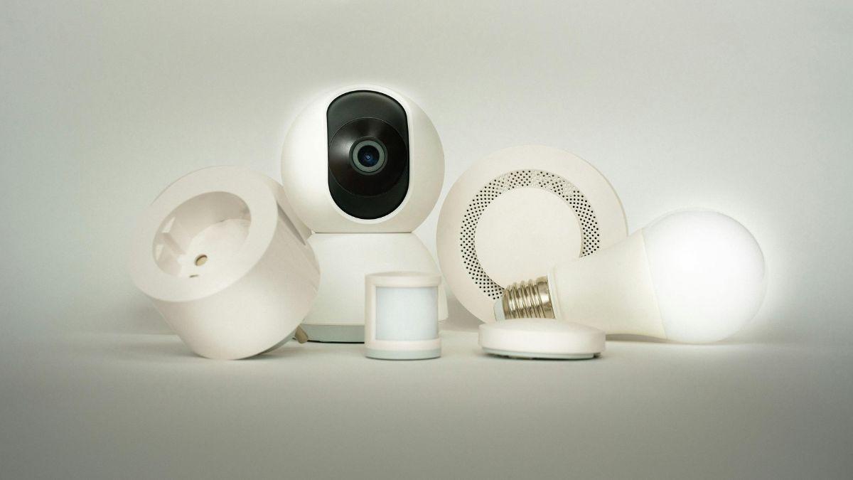 A collection of smart home devices, including a security camera, motion sensor, smart plug, speaker, and smart light bulb, all in white.