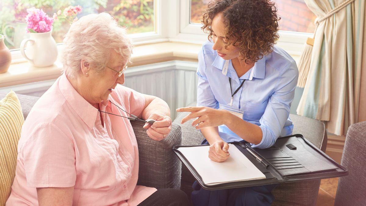 A caregiver in a blue uniform explains a medical alert pendant to an elderly woman sitting on a couch.
