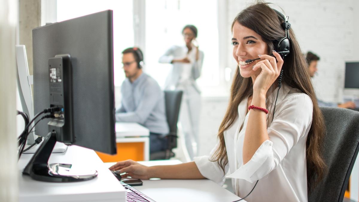 A smiling customer service representative wearing a headset assists a caller in a busy office.