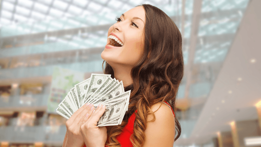 Excited woman holding a fan of dollar bills in her hands, standing inside a shopping mall.