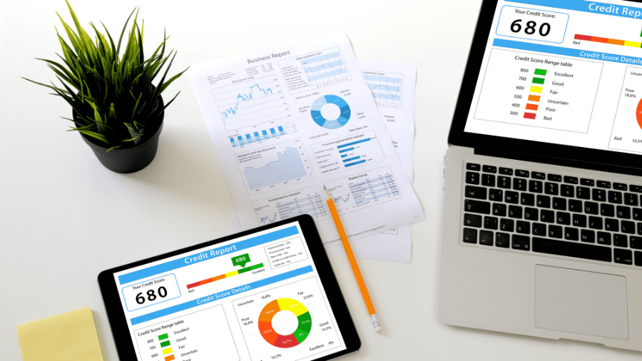 A tablet and a laptop display credit report summaries with a score of 680. Business reports with graphs and charts are scattered on the white desk alongside a pencil, sticky notes, and a small potted plant.