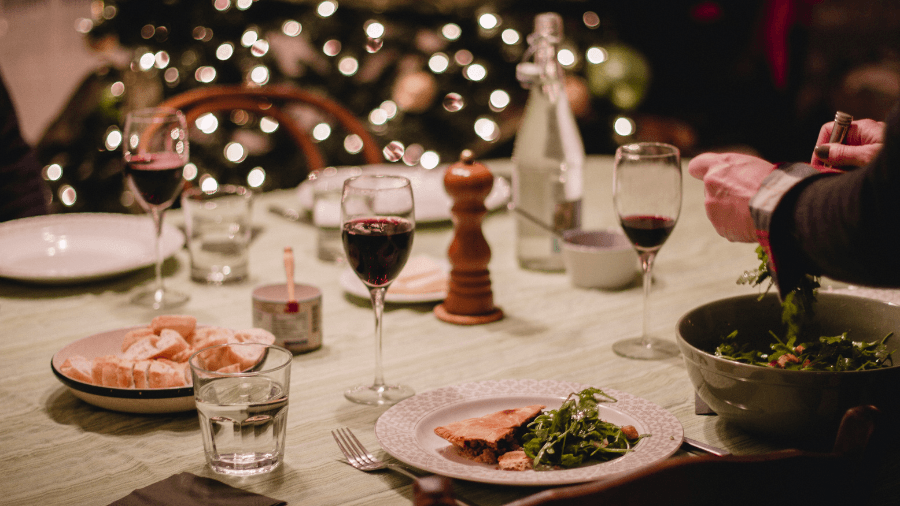Elegant holiday dinner table with red wine glasses, plates of food, and Christmas tree lights in the background.
