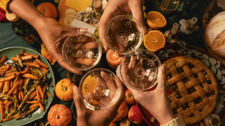 Four hands toasting with white wine glasses over a festive table filled with autumn food and pumpkins