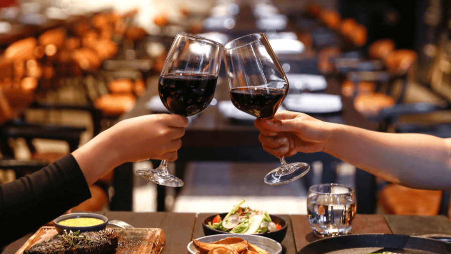 Two people clinking red wine glasses over a dinner table at a restaurant.