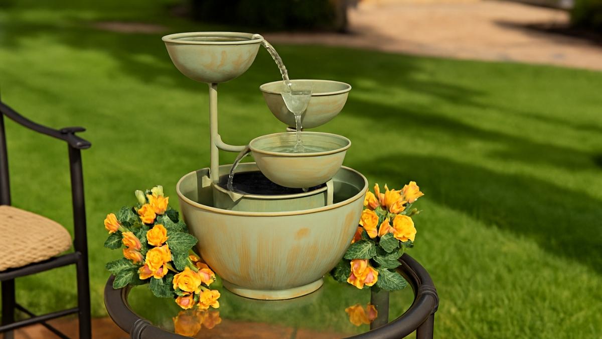 A three-tier decorative metal fountain with flowing water sits on a patio table surrounded by yellow flowers.