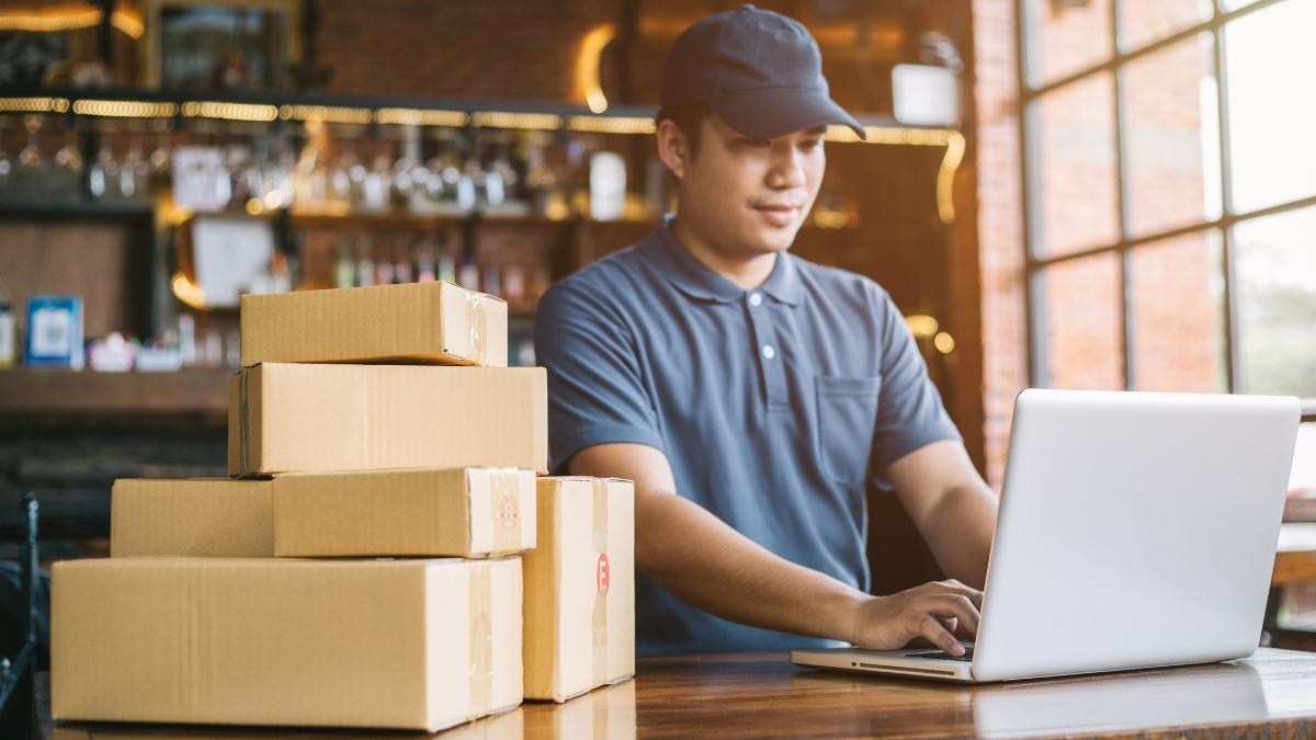 A man in a cap manages Shopify order shipments on a laptop with stacked boxes beside him.