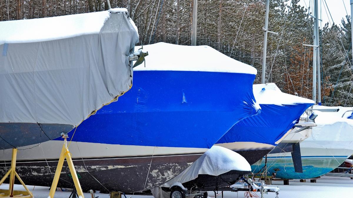Boats covered with protective winter boat covers sit in storage, topped with snow in a marina.