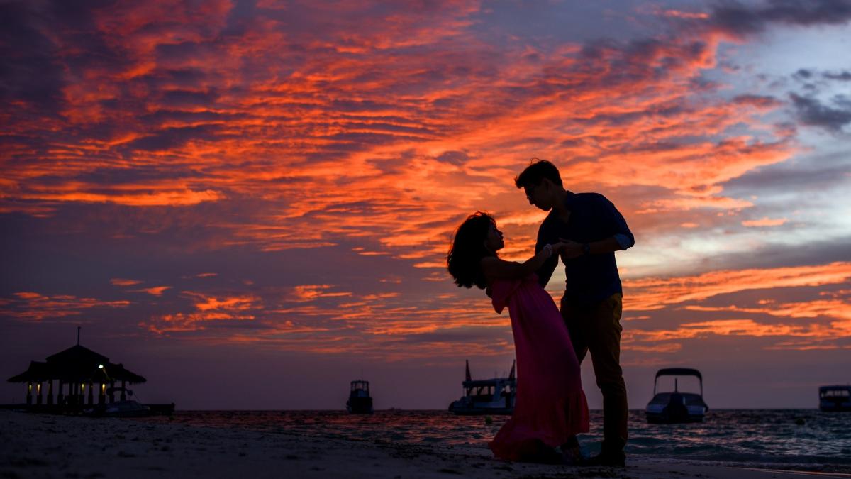 A couple sharing a romantic moment on the beach at sunset, surrounded by vivid orange and purple skies with boats in the background.
