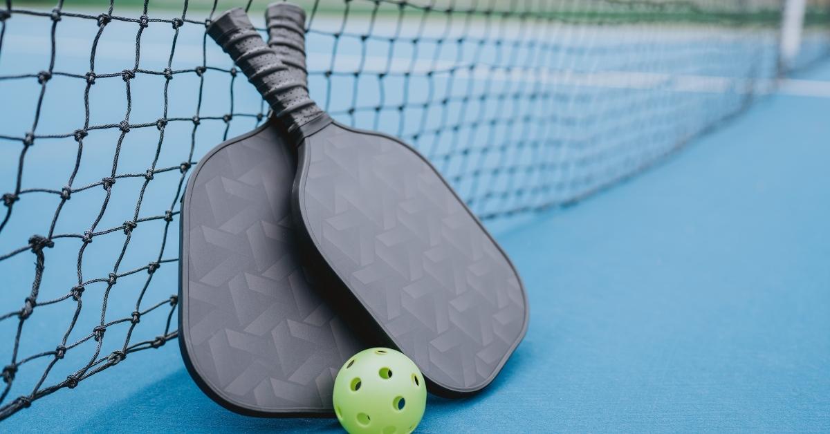 Two pickleball paddles and green ball resting by net on outdoor court surface