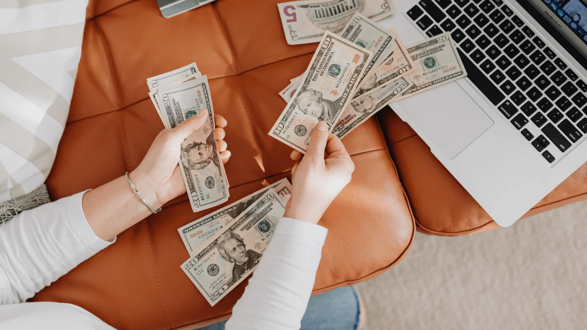 Person counting US dollar bills beside a laptop, representing money management, personal finance, budgeting, and saving money at home.